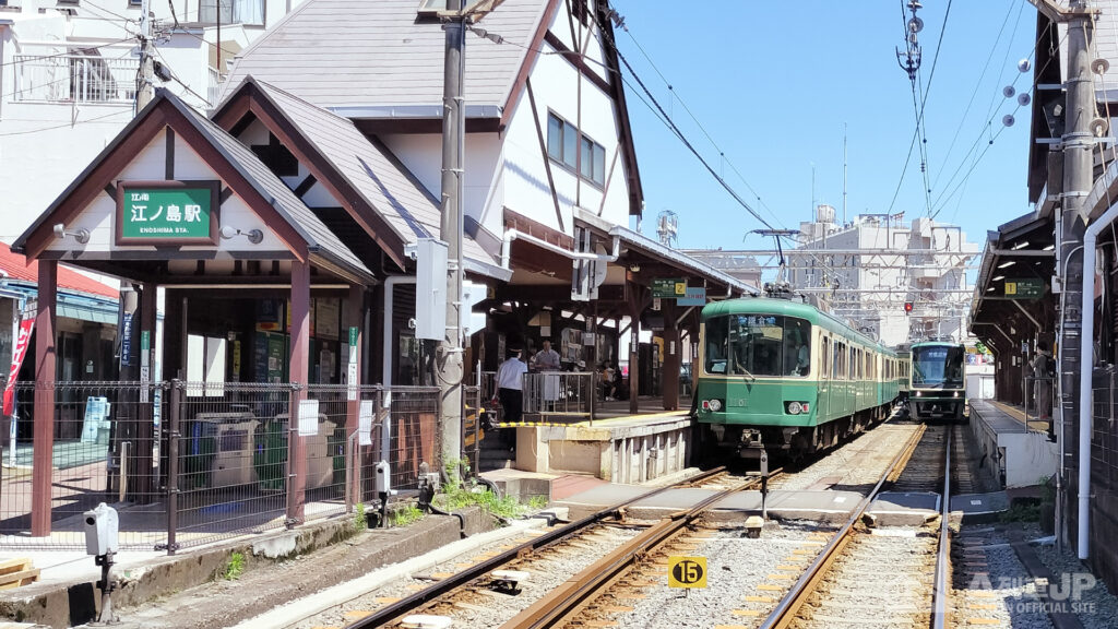 Green commuter train on tracks at Enoshima Station platform in Japan, with passengers waiting under a wooden station canopy and overhead wires visible.