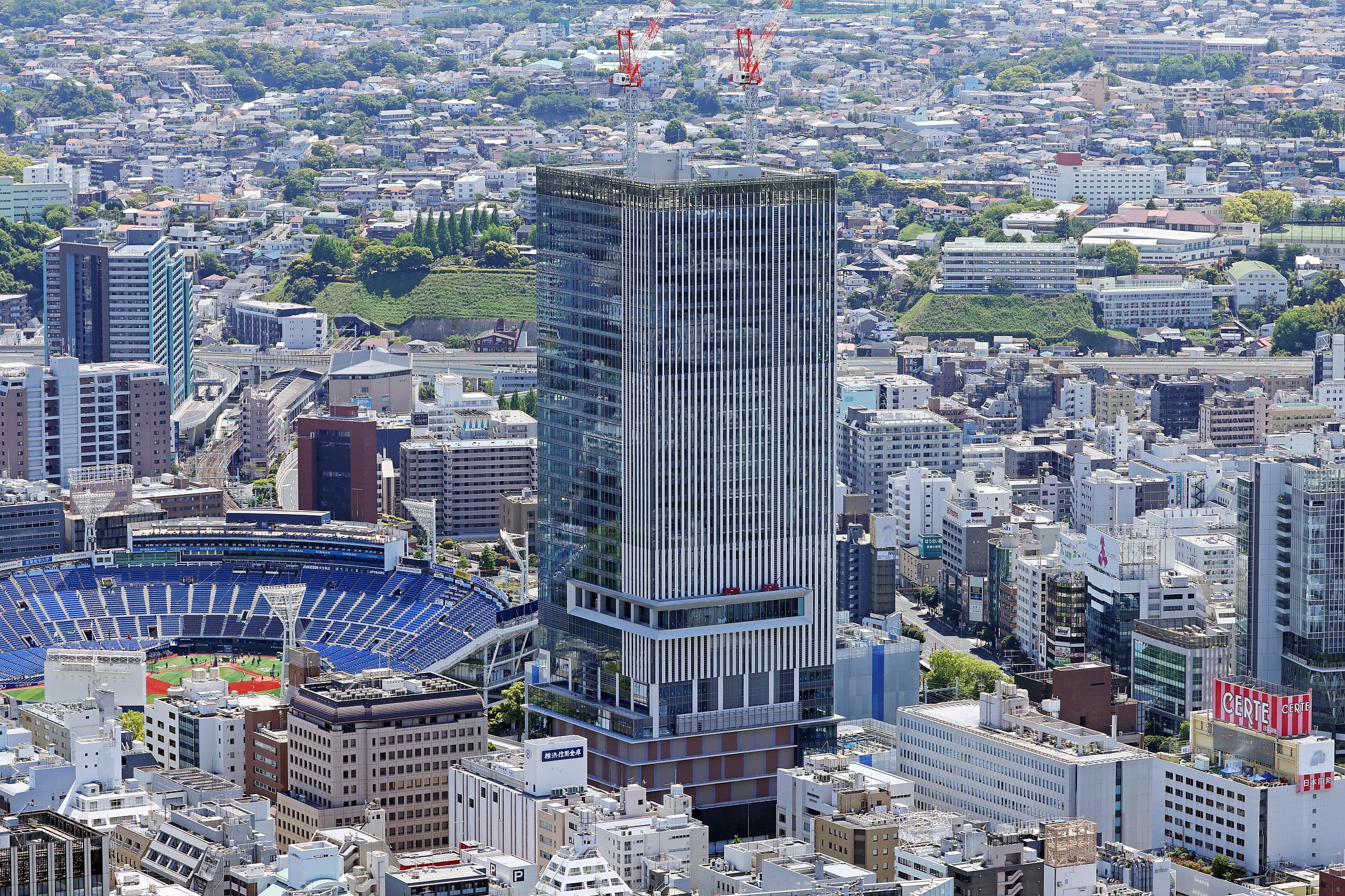 Aerial view of a dense city with a tall glass skyscraper under construction, two red cranes on top, and a blue stadium at street level.