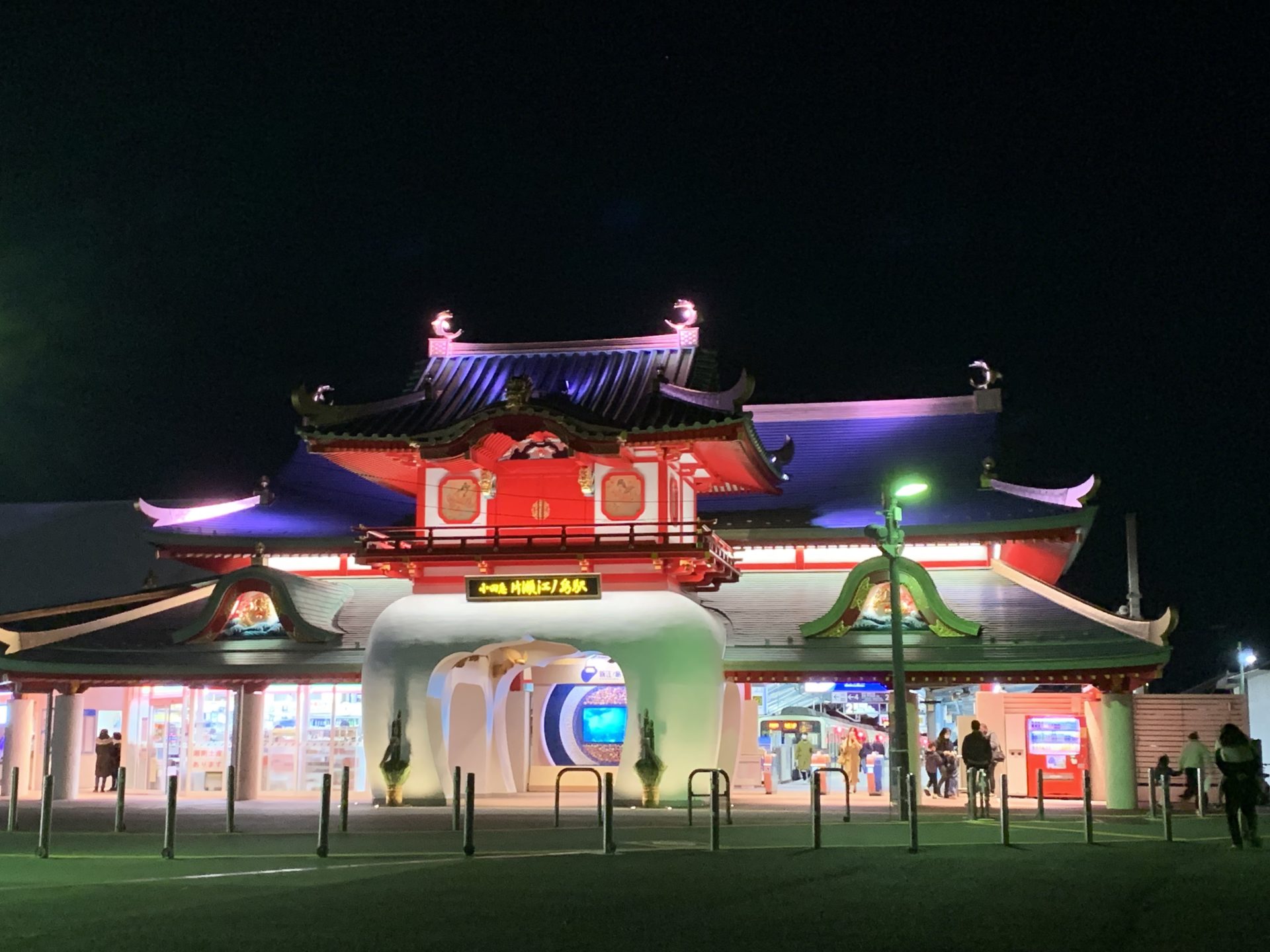 Night view of a brightly lit Asian temple facade with red and green neon lights, and people near the entrance and market stalls in front.