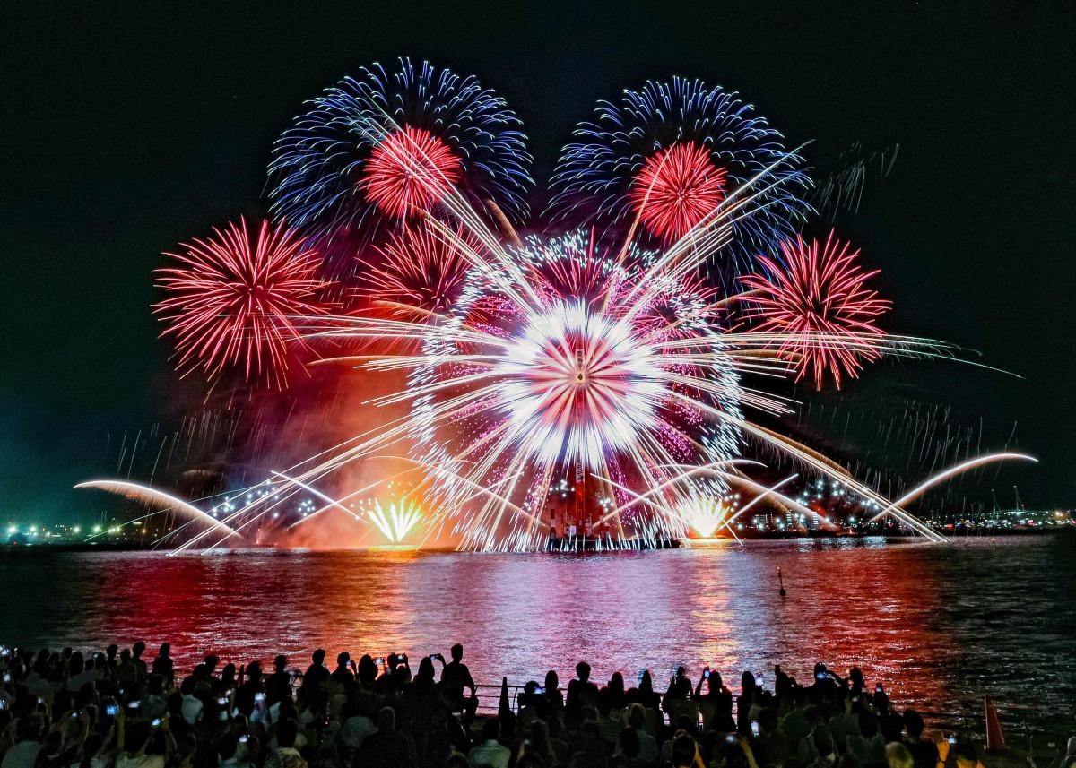Night scene of colorful fireworks exploding over a waterfront, with a silhouetted crowd watching and reflections on the water.
