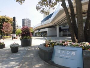 Sunny urban plaza outside an elevated walkway near Yokohama Park, with flower planters and a sign reading Yokohama Park.