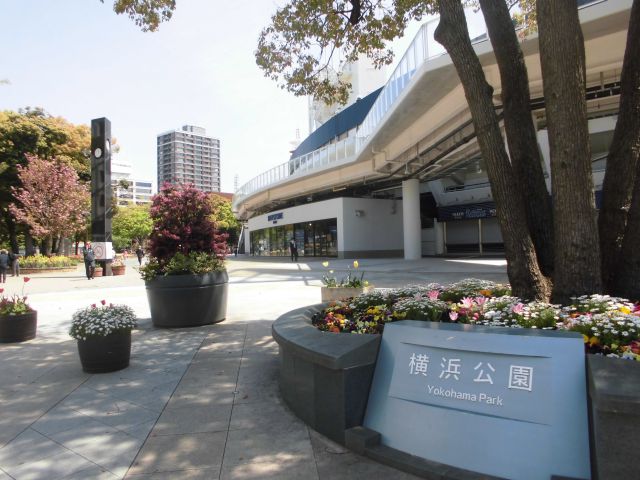 Sunny urban plaza outside an elevated walkway near Yokohama Park, with flower planters and a sign reading Yokohama Park.