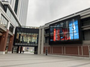 Modern urban plaza with glass-fronted buildings and elevated walkway connecting structures, overcast sky above.