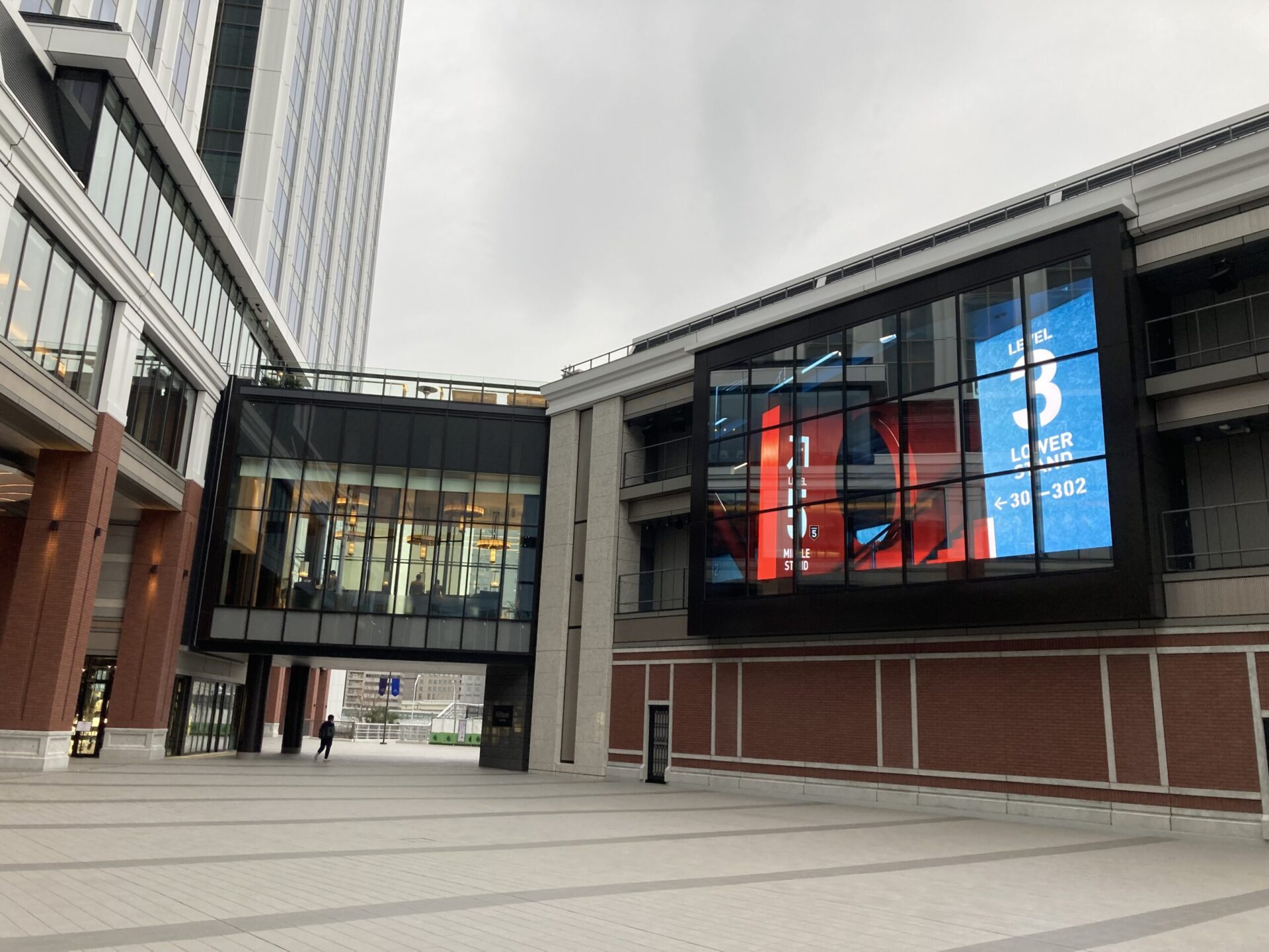 Modern urban plaza with glass-fronted buildings and elevated walkway connecting structures, overcast sky above.