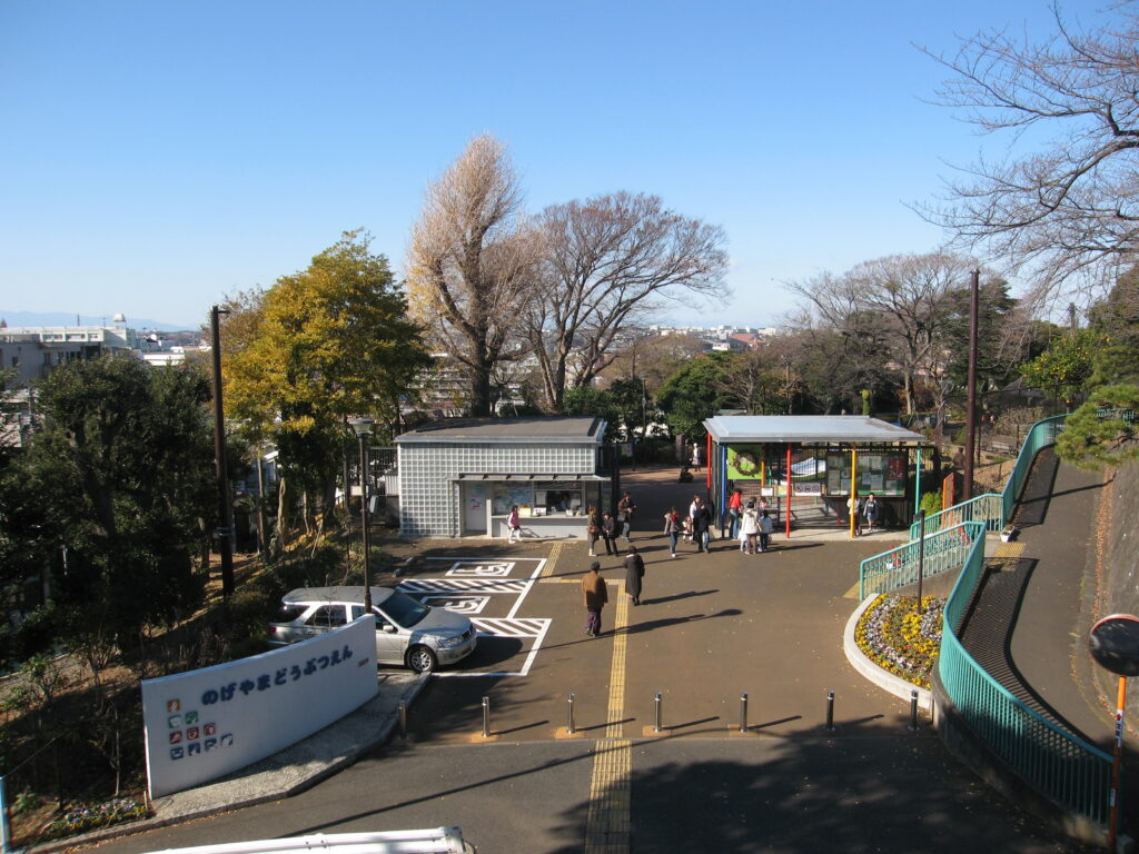 Park entrance and walkway with people strolling under a clear blue sky, bare trees and city buildings in the distance.