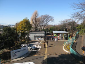 Park entrance and walkway with people strolling under a clear blue sky, bare trees and city buildings in the distance.