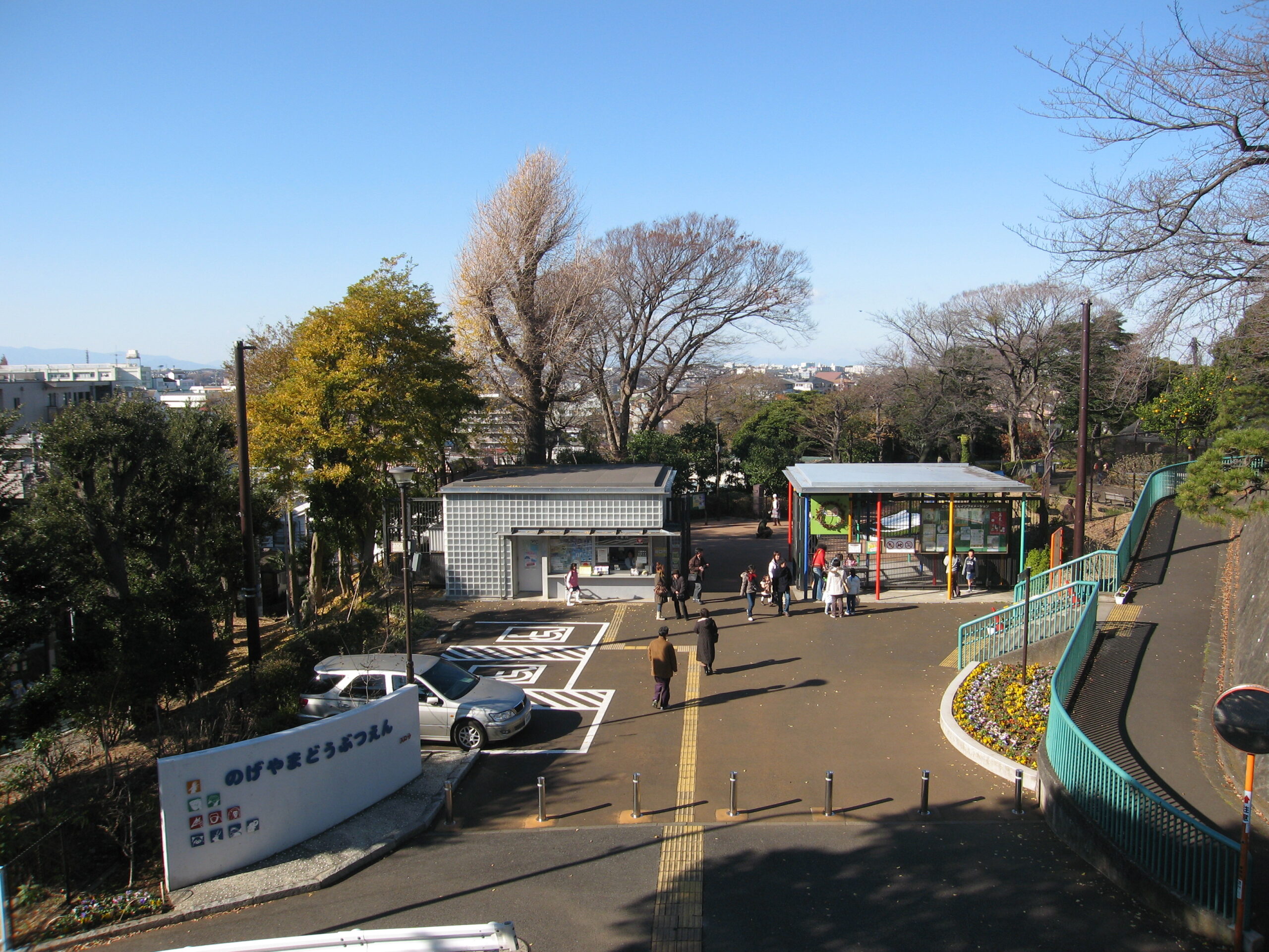 Park entrance and walkway with people strolling under a clear blue sky, bare trees and city buildings in the distance.