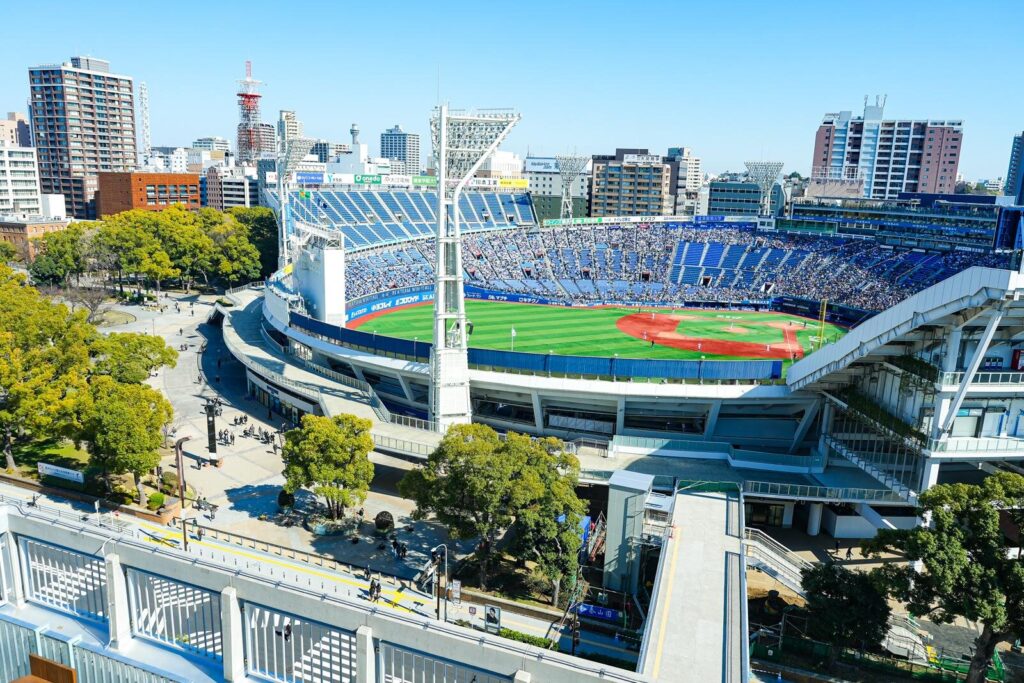 Overhead view of a baseball stadium with blue seating, green field, and a clear blue sky behind a city skyline.
