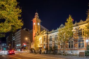Nighttime street scene with a red brick clock-tower building illuminated beside a tree-lined avenue and passing cars on the road