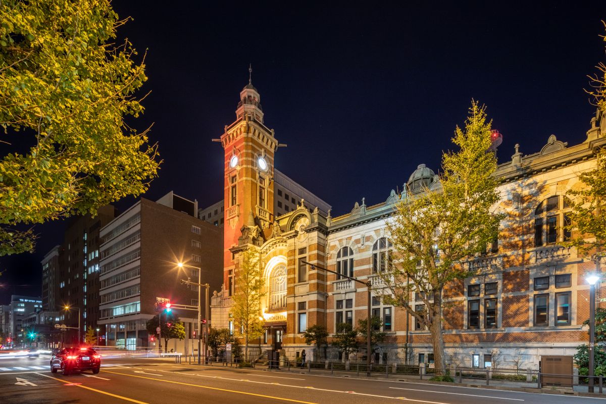 Nighttime street scene with a red brick clock-tower building illuminated beside a tree-lined avenue and passing cars on the road