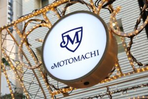 Circular illuminated storefront sign with blue Motomachi emblem and 'MOTOMACHI' text, framed by warm string lights