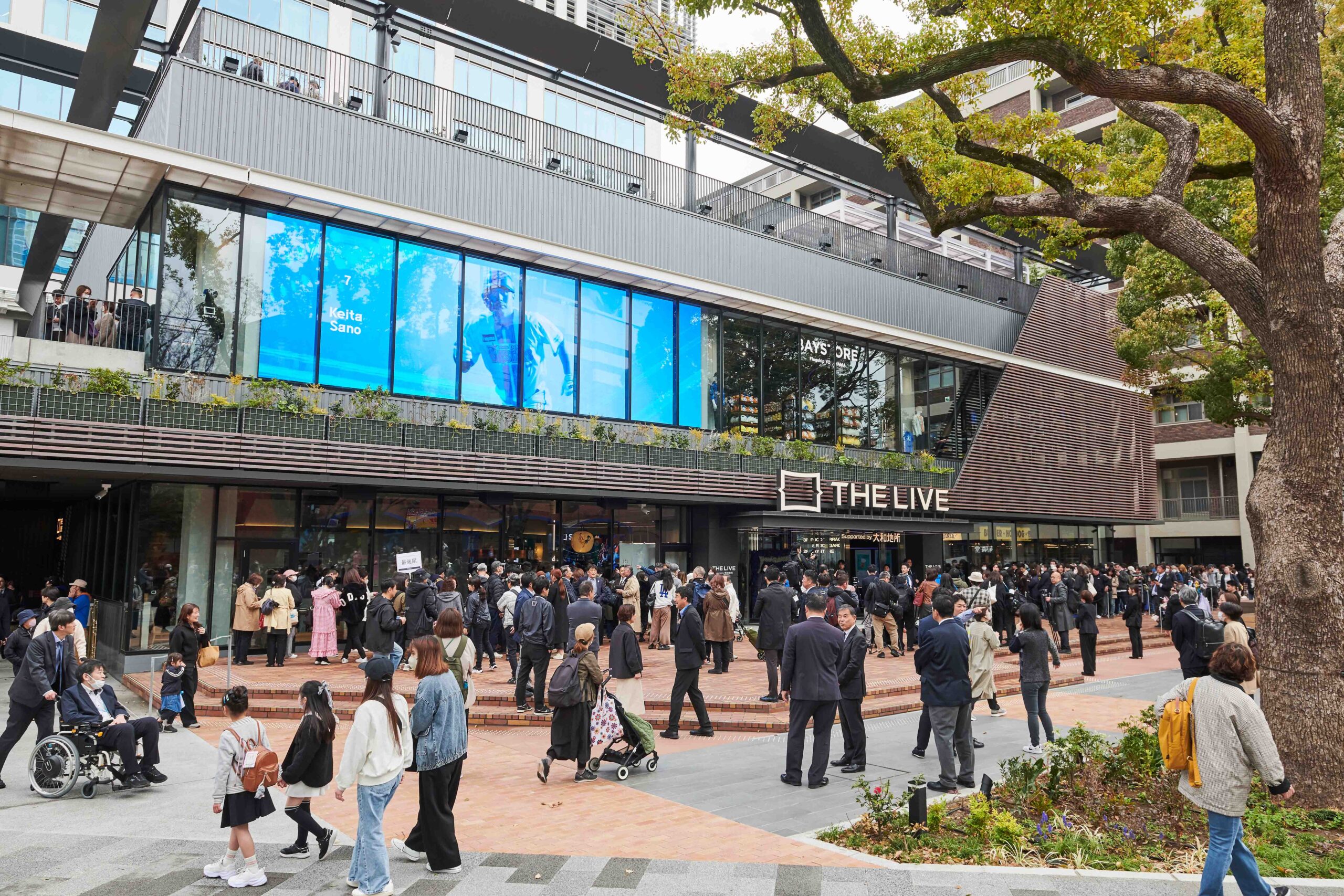 Crowd of people gathers outside a modern glass-front building with blue storefront displays and greenery nearby.