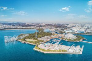 Aerial view of a waterfront island park with multiple buildings, blue water, and a bridge to the mainland under a clear sky.