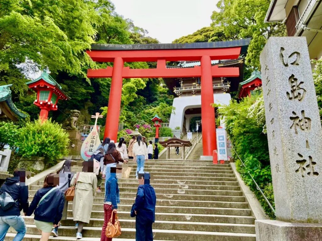Visitors ascend stone steps beneath a bright red torii gate at a Japanese shrine surrounded by greenery.