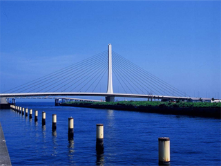 Cable-stayed bridge with a tall white pylon and radial cables over a blue river, viewed from the water's edge.