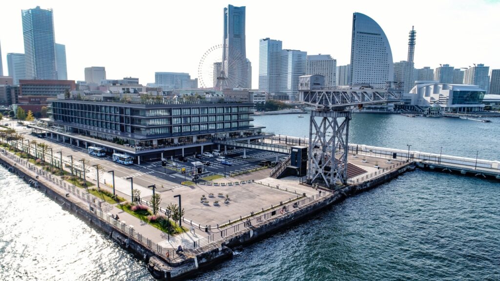 Aerial view of a modern waterfront district with a multi‑story glass building, a ferris wheel, and a large metal crane over the harbor.