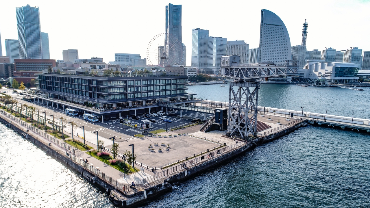 Aerial view of a modern waterfront district with a multi‑story glass building, a ferris wheel, and a large metal crane over the harbor.