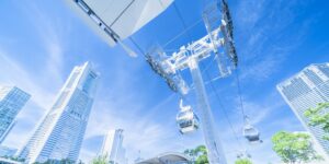 Cable car gondolas suspended from a white tower over a modern city with tall glass buildings under a clear blue sky.