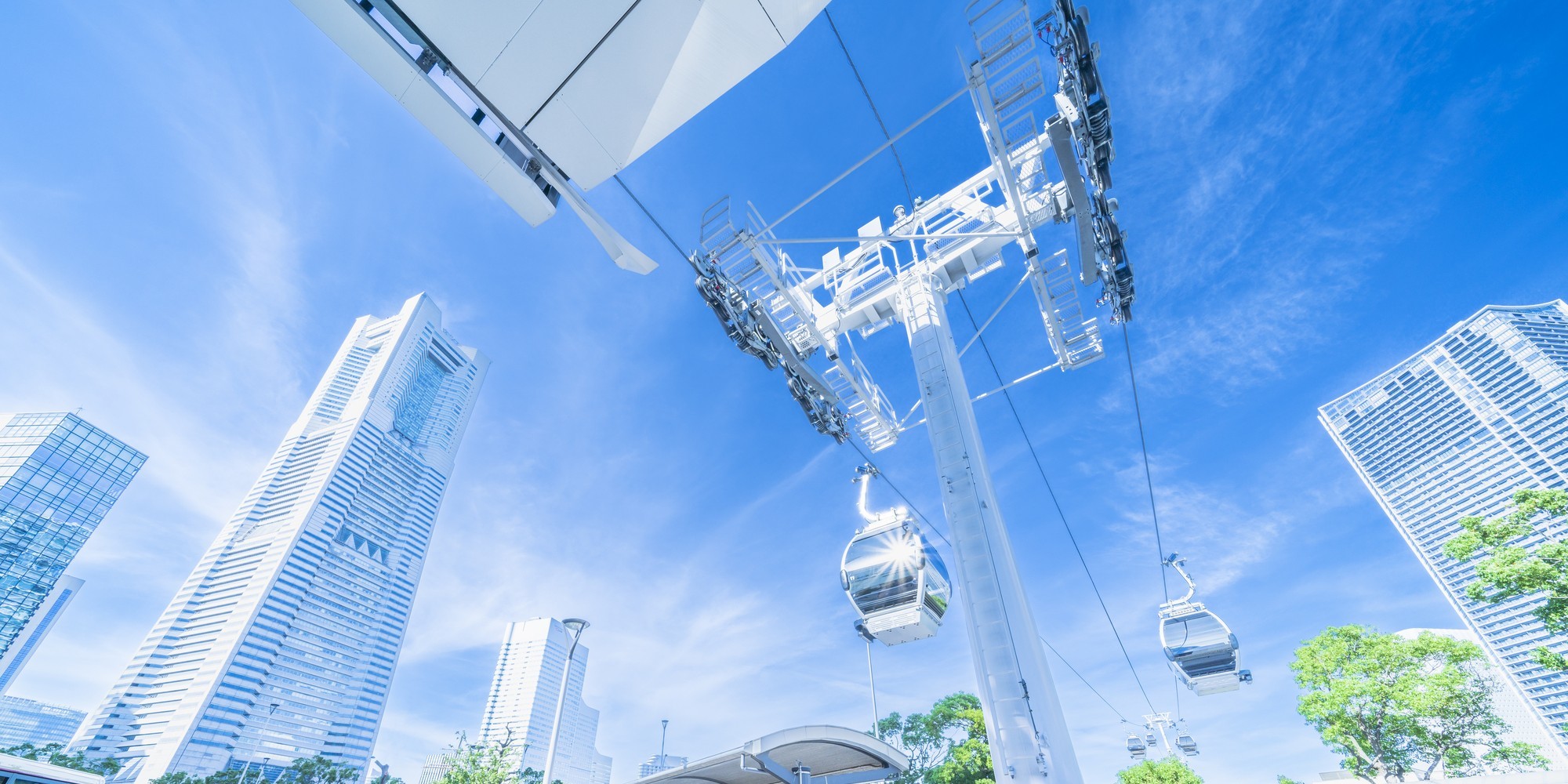 Cable car gondolas suspended from a white tower over a modern city with tall glass buildings under a clear blue sky.